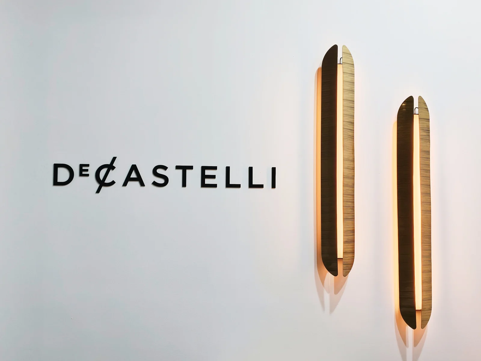 Логотип DE CASTELLI и вертикальные бра на белой стене